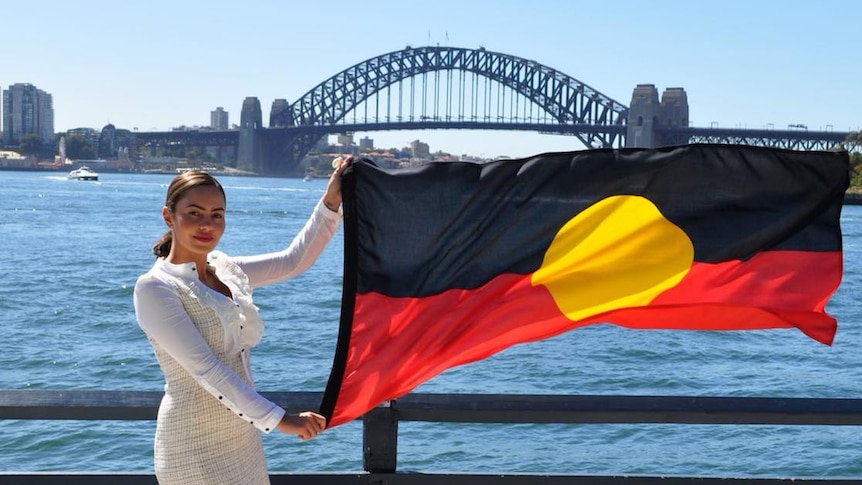 Woman stands on Sydney harbour with Aboriginal flag waving in her hands. She is in front of the Sydney Harbour Bridge