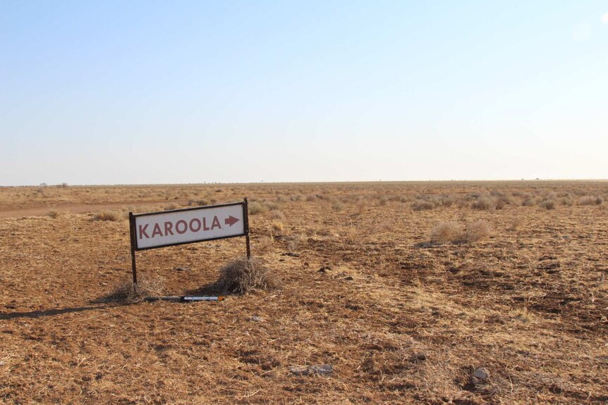 A sign reading "Karoola" with an arrow pointing right against a flat plain of dry grassy tufts.