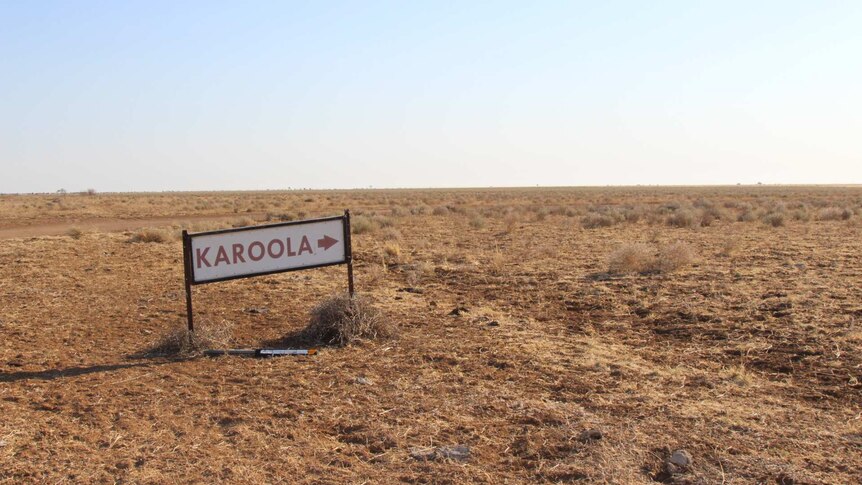 A sign reading "Karoola" with an arrow pointing right against a flat plain of dry grassy tufts.