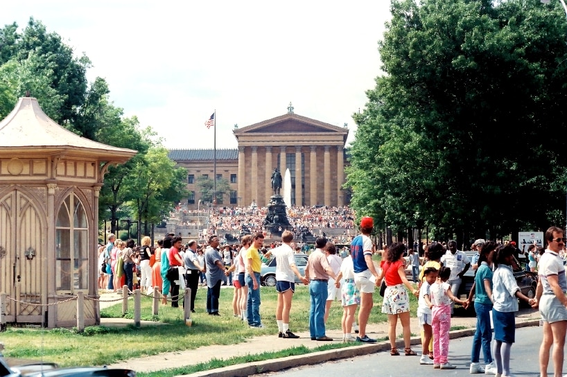 A crowd of several dozen people join hands in a line with a building in the background