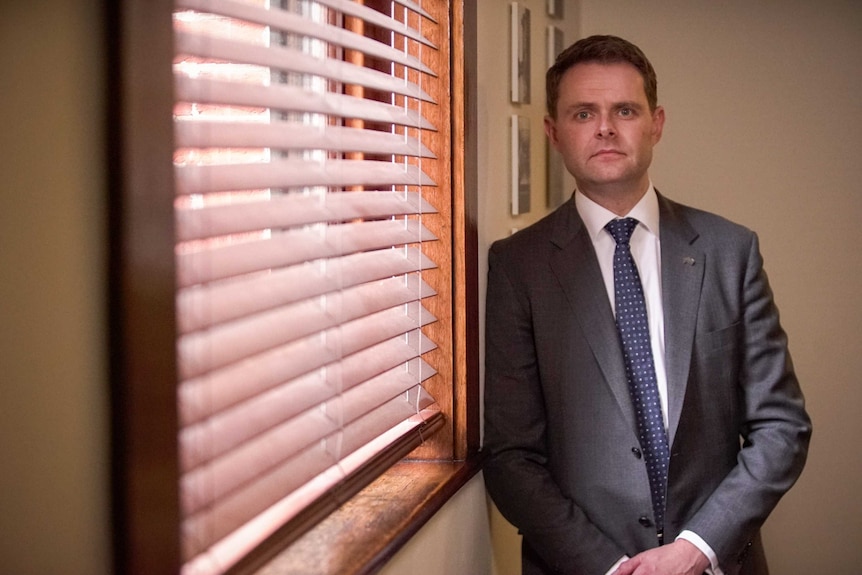 A man wearing a suit next to a window with blinds