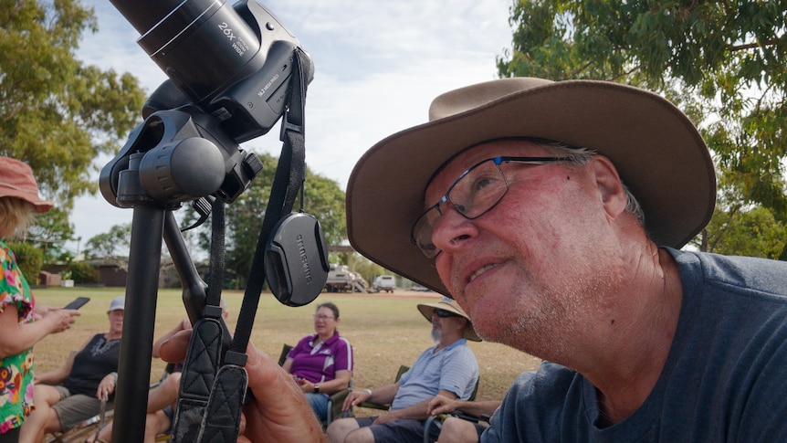 A man wearing glasses and a hat angles his camera towards the sky and looks through the viewfinder.