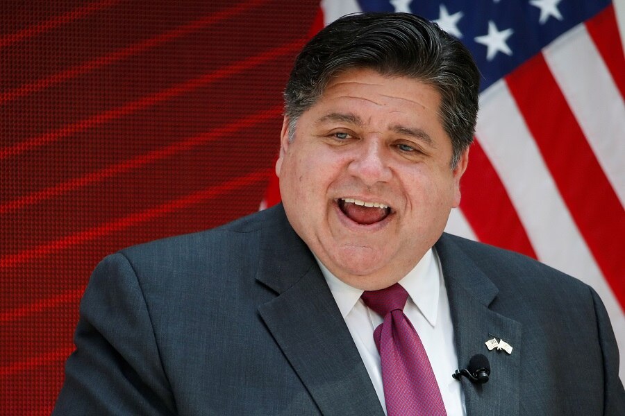 A middle-aged white man with dark hair in a suit stands in front of an American flag.