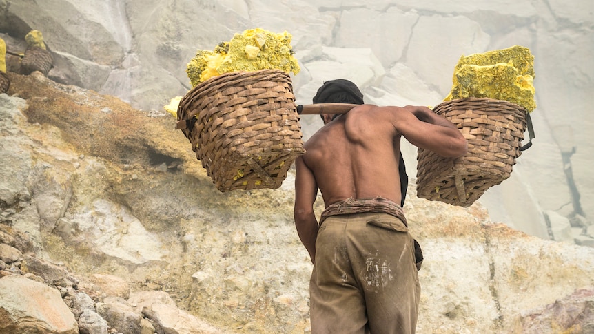 back of man carrying yellow blocks of sulfur in baskets on his back up rocky landscape with tattered pants and no shirt