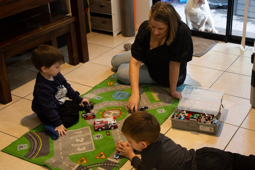 Woman sits on the floor with two young boys, playing with toy cars.