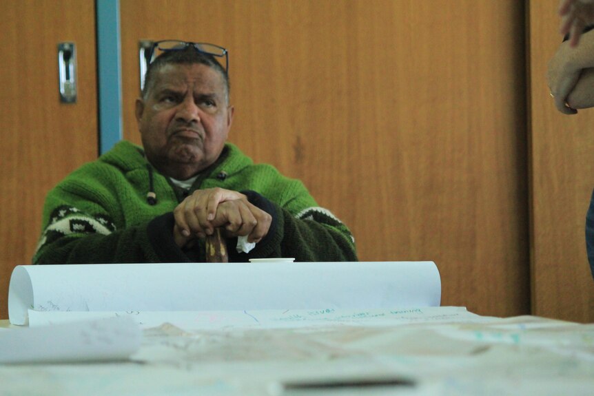 An older indigenous man with a green jumper sits at a table, with a serious expression.