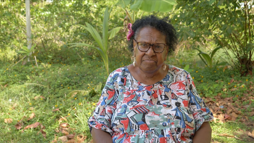 Aunty McRose Elu standing infront of tropical greenery with a bright shirt on wearing glasses and looking serious