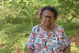 Aunty McRose Elu standing infront of tropical greenery with a bright shirt on wearing glasses and looking serious