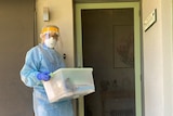 A woman in full PPE is holding a tub of supplies, standing outside the front door of a house.