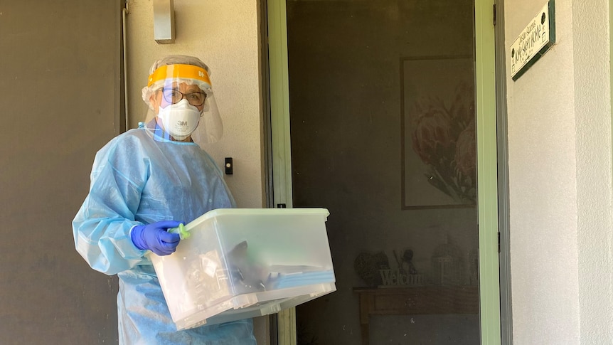 A woman in full PPE is holding a tub of supplies, standing outside the front door of a house.