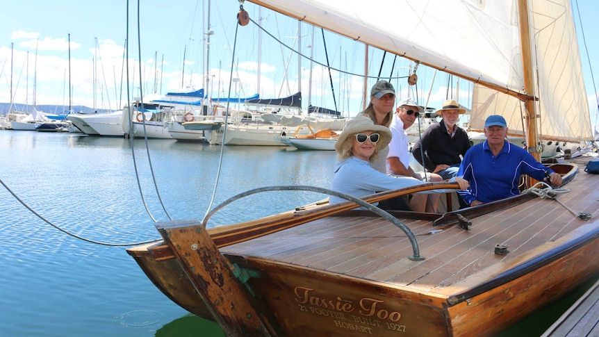 Five people sit inside a wooden boat and smile. The boat is floating in a marina on a picturesque Tasmanian day