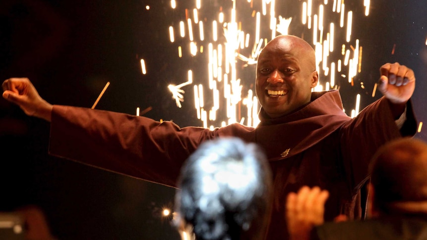 Peter Tabichi smiles and holds his arms up as people in the foreground clap.