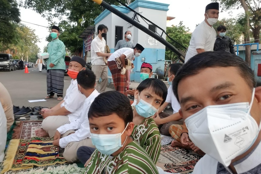 A man wearing a mask with other people in the background also wearing masks waiting to pray