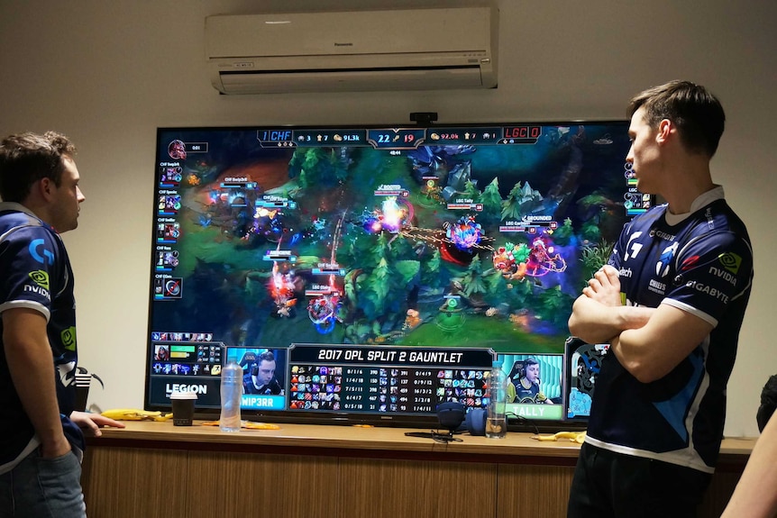 Three players from The Chiefs looking at a big wide screen of a League of Legends game.
