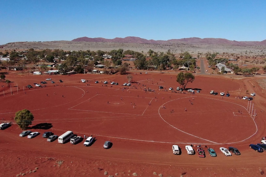 A red dirt football oval surrounded by cars with trees and a small community in the mid-distance