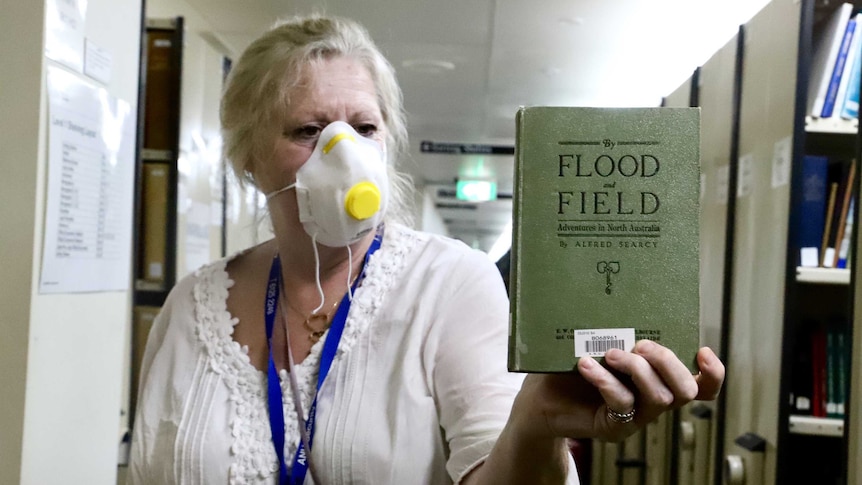 A woman wearing a mask holds a book titled 'By Flood and Field' in a library.