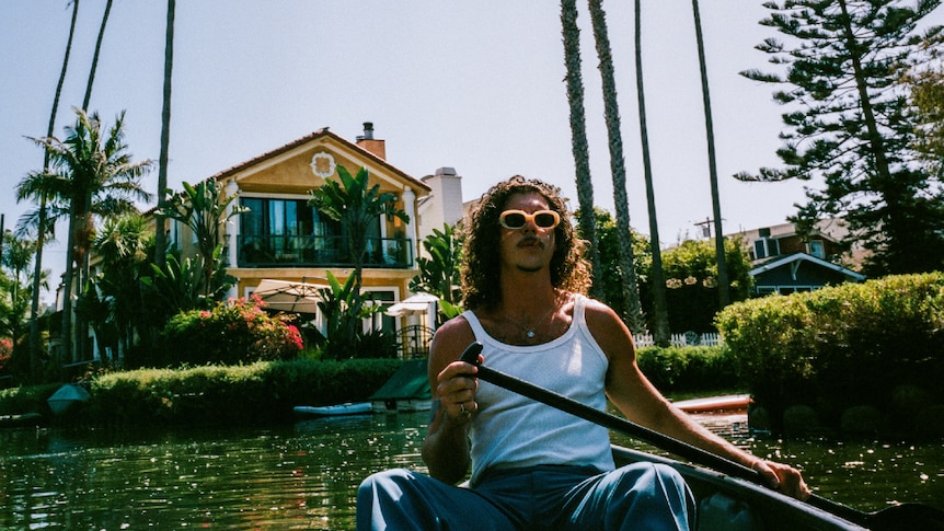 Keli Holiday (Adam Hyde from Peking Duk) in a boat, holding an oar. Behind him is a lavish house on the water and palm trees