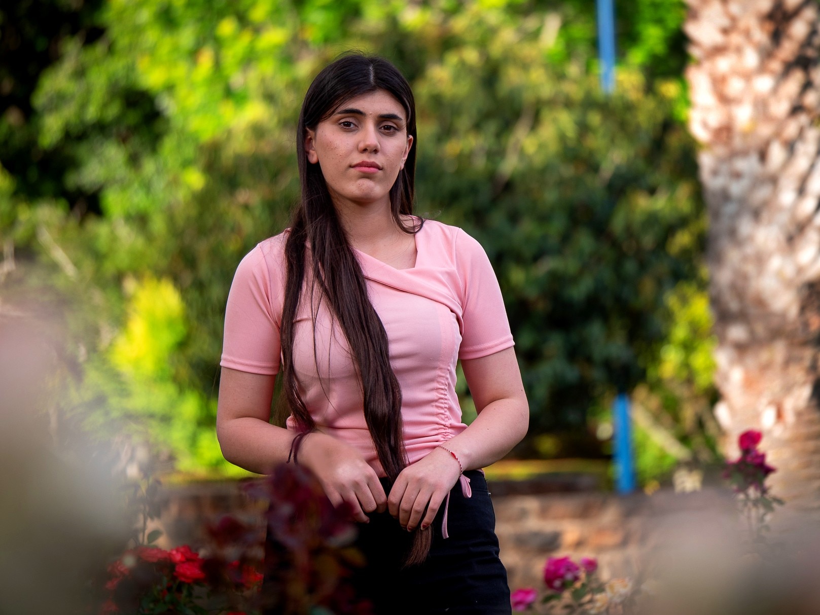 A woman with long black hair in a pink shirt. Standing near roses with green trees in the background.