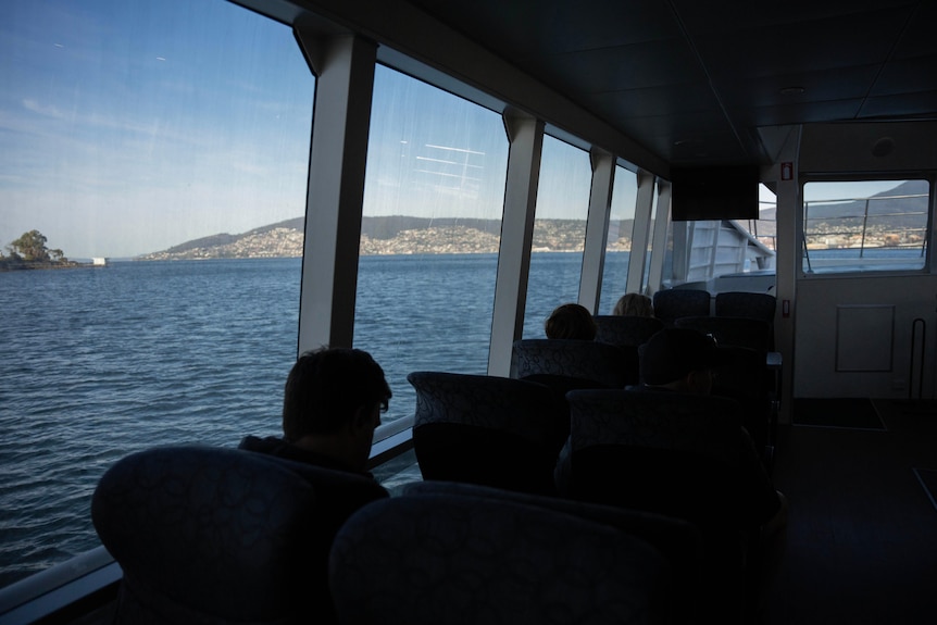 A silhouette of several passengers against ferry windows with the River Derwent and Hobart suburbs in the background.