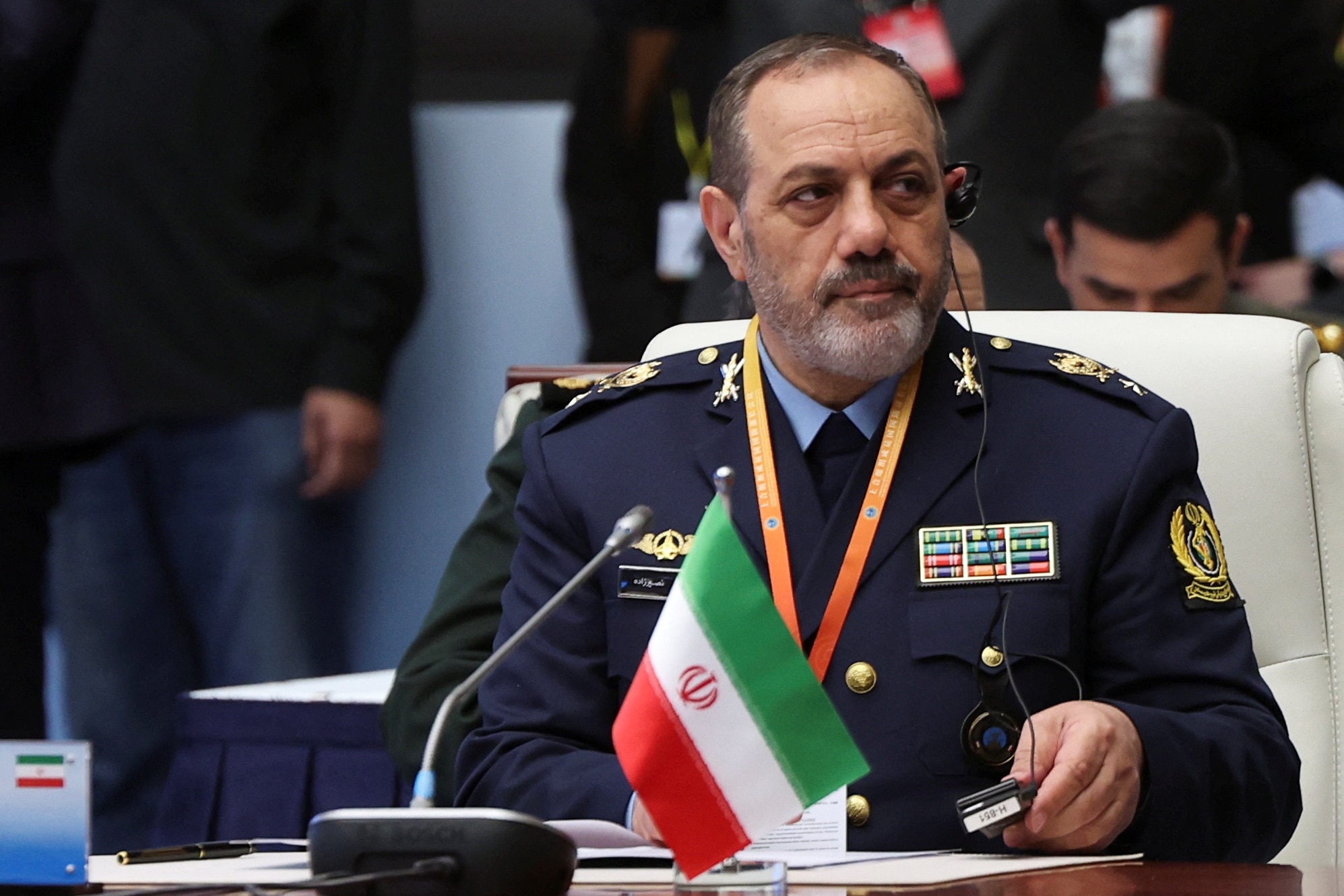 Aziz Nasirzadeh wearing a navy military uniform sits at a desk with an Iranian flag, looking off to his side.