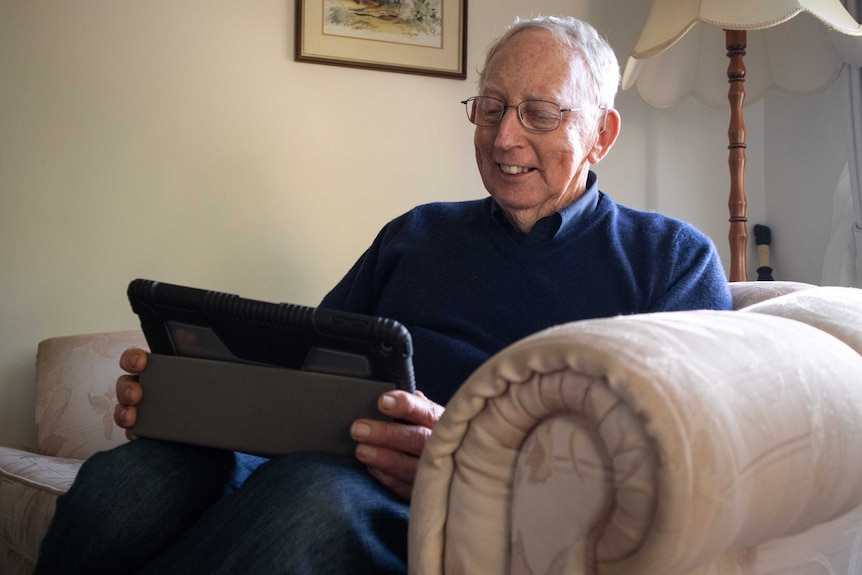 John Broadbent sitting in armchair looking at his iPad.