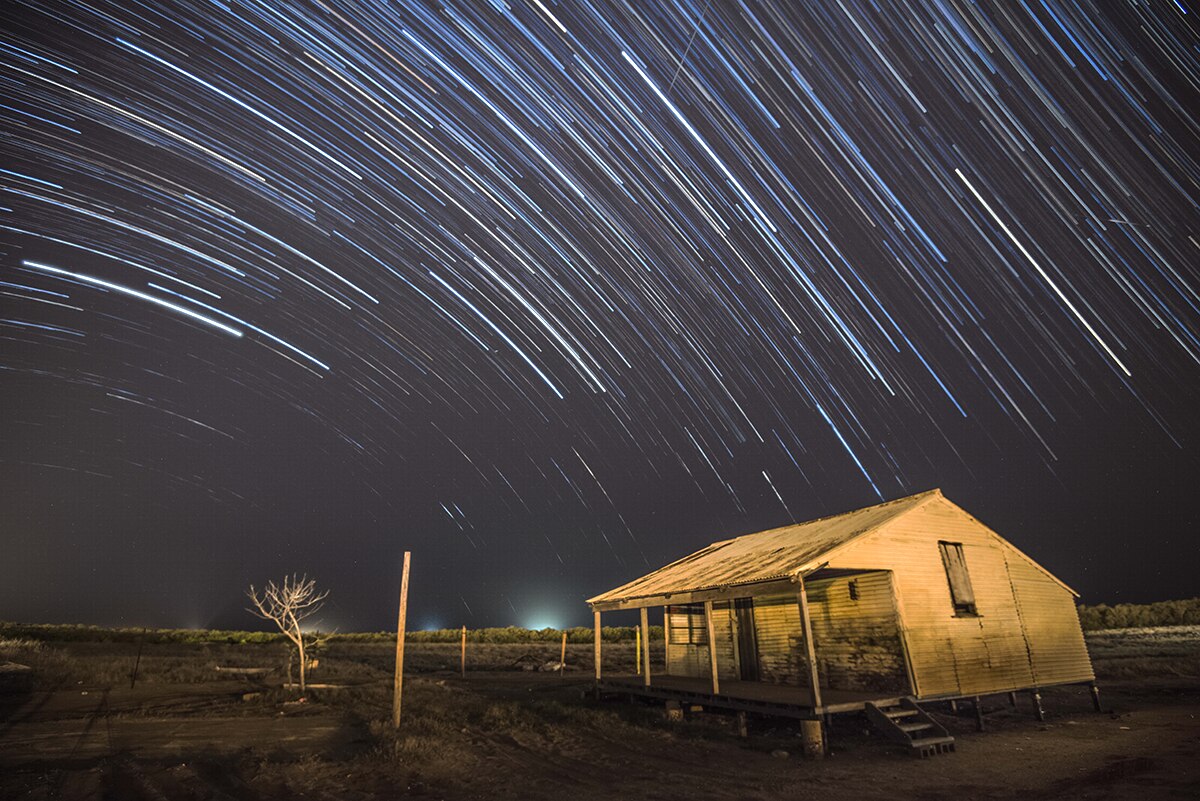 A long exposure leaves star trails in the night sky above a shed in a field on a rural property.