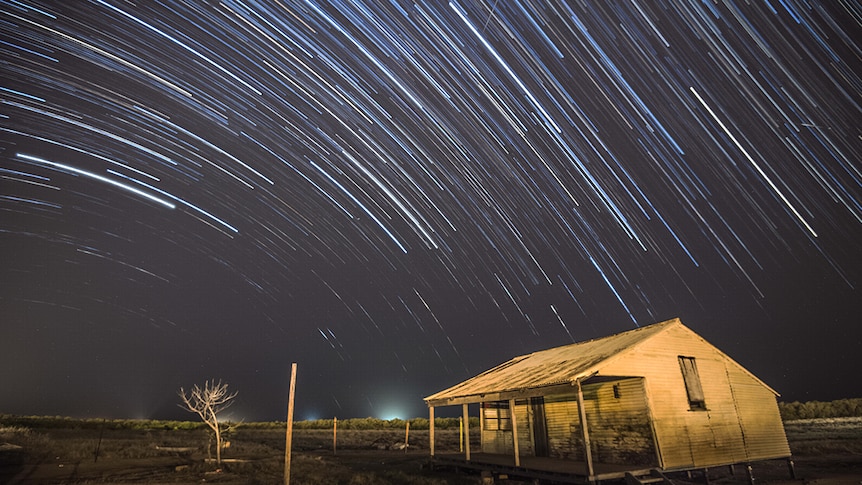 A long exposure leaves star trails in the night sky above a shed in a field on a rural property.