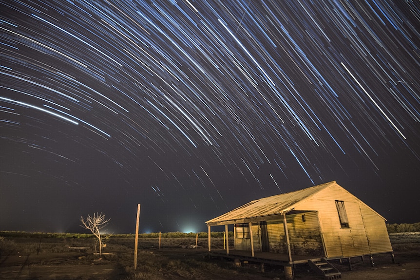 A long exposure leaves star trails in the night sky above a shed in a field on a rural property.