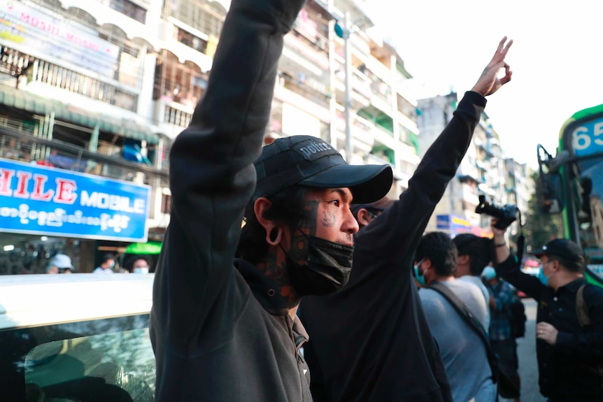 Two men, one wearing face mask and cap in front of photo, raise three fingers to air.