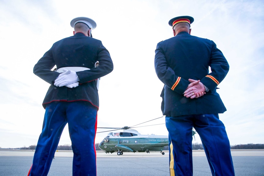 Two US marines in dress uniform stand with their backs turned to the camera, looking at the Marine One helicopter