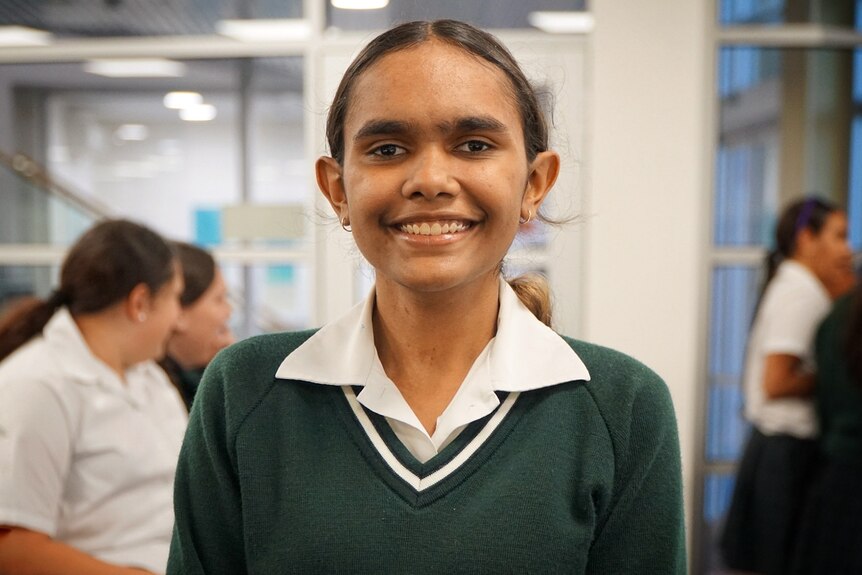 A young Aboriginal girl wearing a white collared shirt under a green sweater smiling broadly at the camera