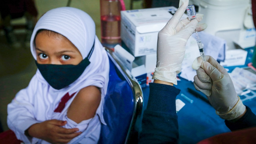 A little girl in a white hijab and black face mask exposes her shoulder behind a pair of gloved hands holding a needle