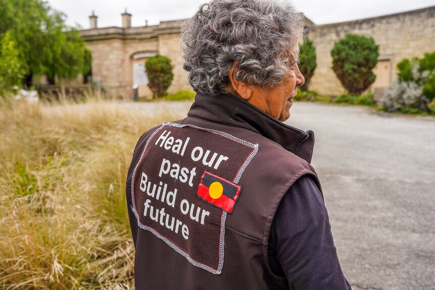 A woman with grey hair, standing wearing a jack vest with the words 'Heal our past, build our future' on the back.