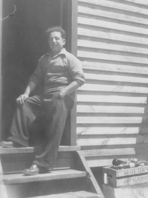 black and white photo of man shirt sleeves rolled up foot on step leaning against weatherboard hut