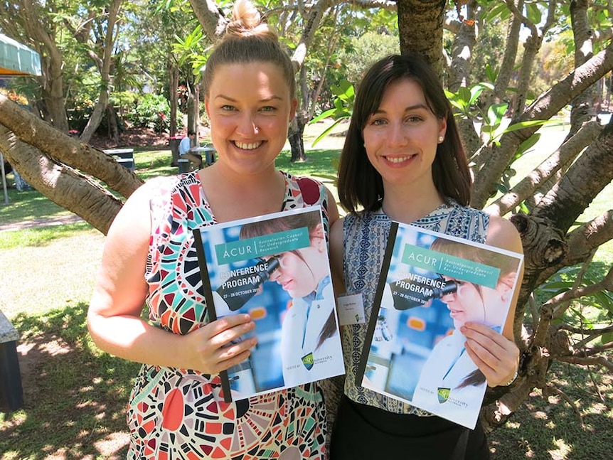 Two women stand in the shade of a tree holding a booklet each titled 'conference program'