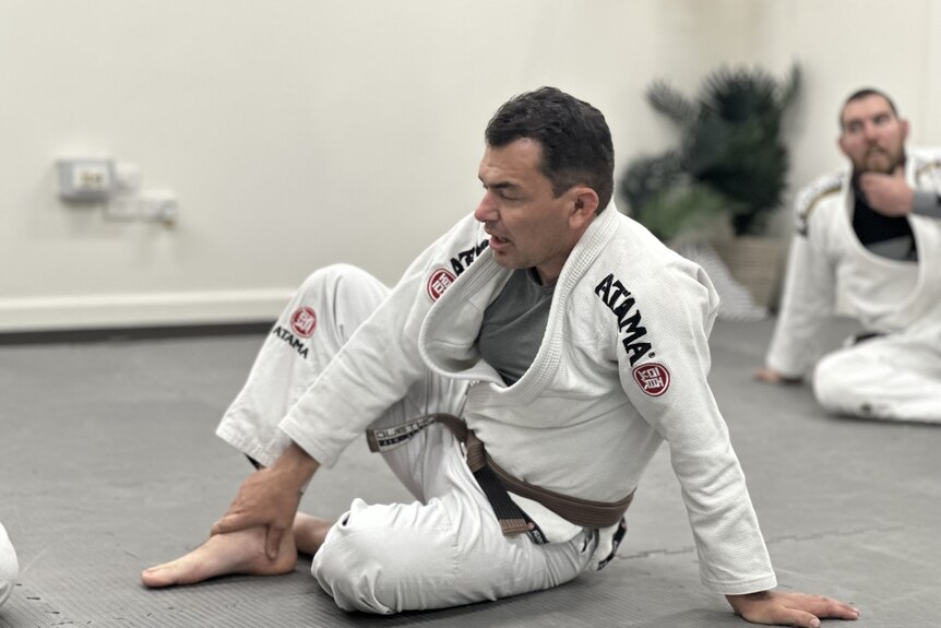 A man sits on a grey mat wearing a white gi and brown ju jitsu belt. 