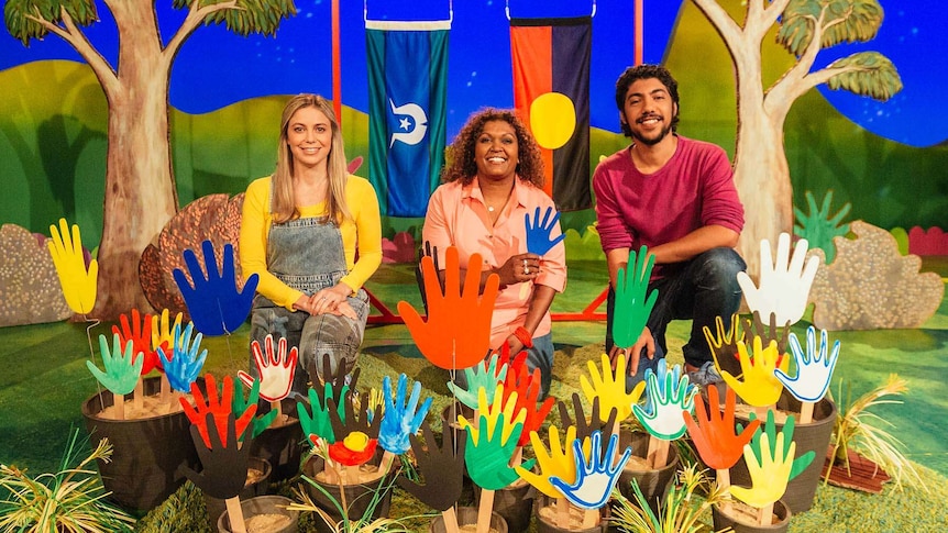 Hunter, Rachael & Emma Donovan behind coloured hand cut outs and the Torres Strait Islander flag and Aboriginal flag behind them