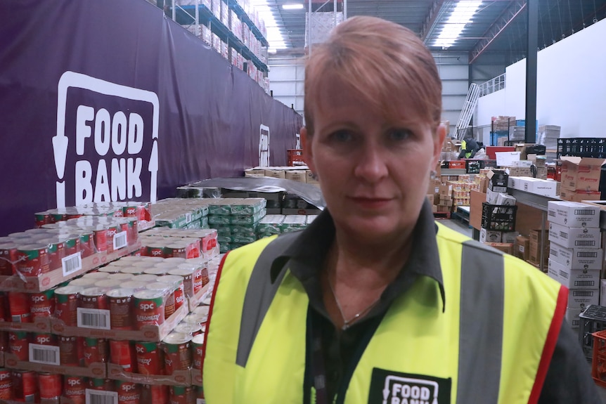 Headshot of woman slightly smiling in Foodbank distribution centre full of cans, wears fluoro vest, large Foodbank sign on wall.