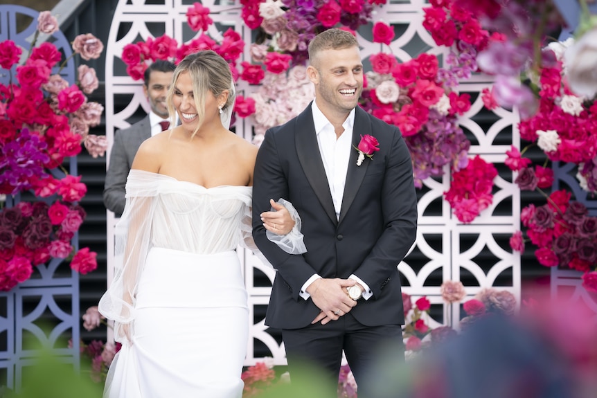 A bride and groom in front of a pink and purple floral arrangement. 