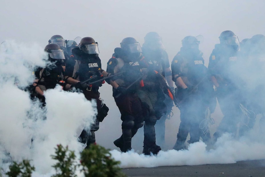 A group of heavily armed police emerge from a cloud of smoke.