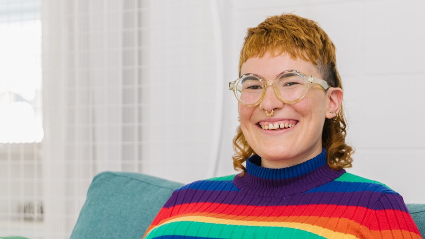 Smiling person wearing eyeglasses and a rainbow striped-jumper sitting on a couch