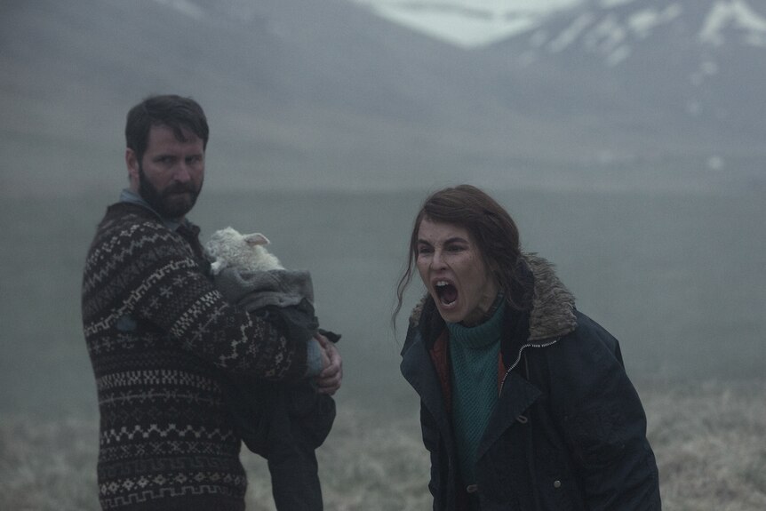 Bearded man in a patterned knitted jumper clutches a lamb to his chest while dark-haired woman in rugged blue jacket yells.