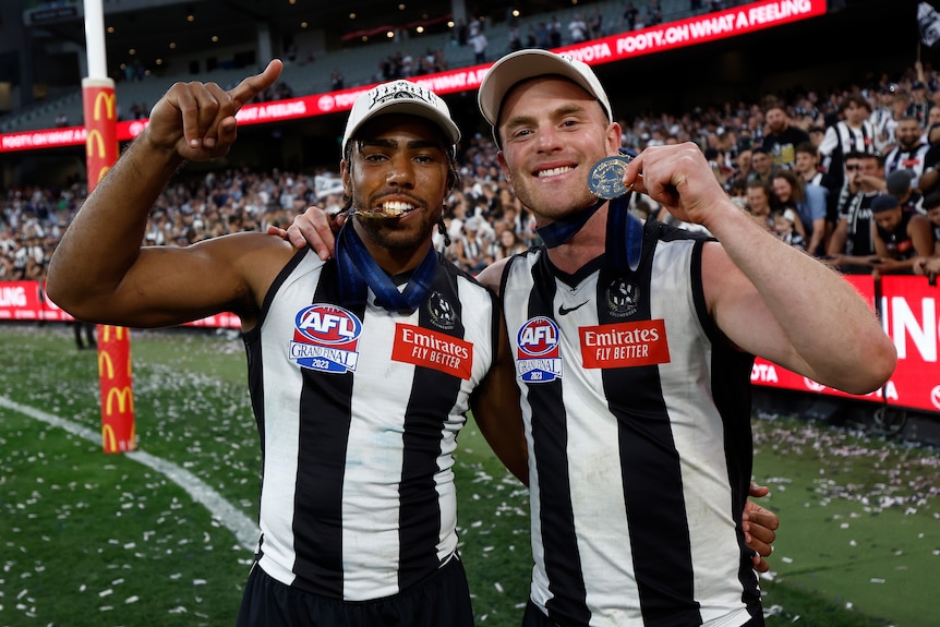 Two cap-wearing Collingwood AFL players smile at the camera as they hold their medals after a grand final win.