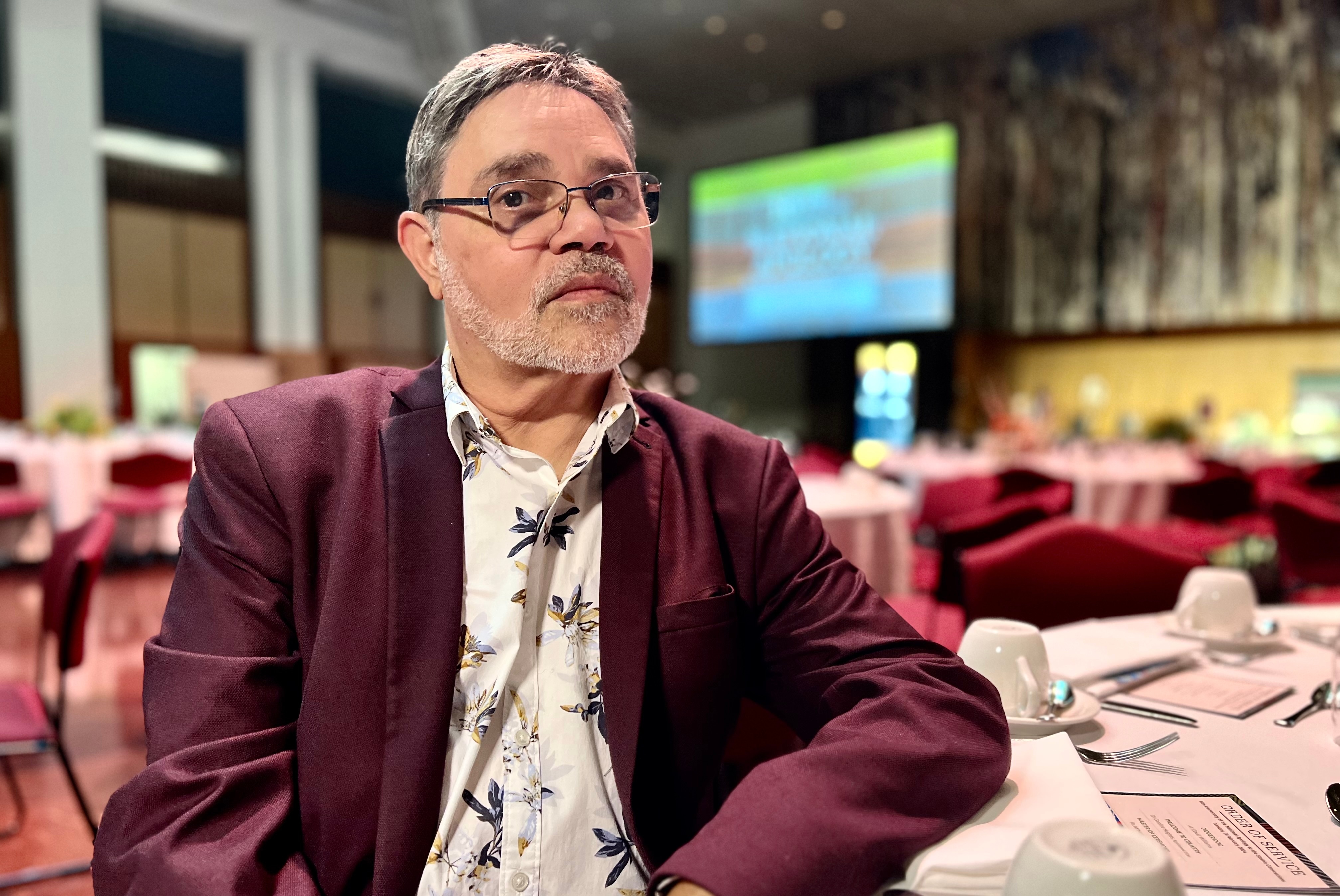 Michael sits at the table in a burgundy suit jacket and floral shirt