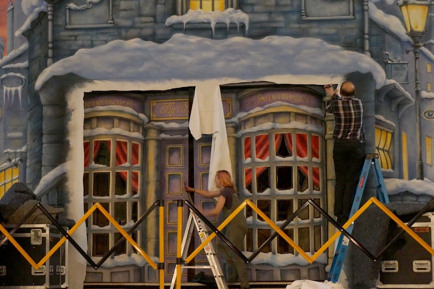 A theatre set of a toy store in wintry 1830's London. Phil Daniels stands on a ladder and trims the painted cloth with scissors.