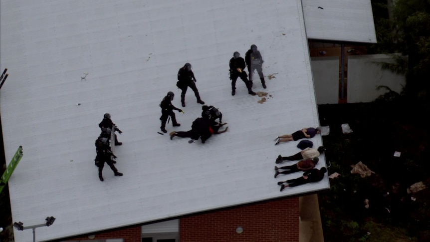 Multiple officers in riot gear making an arrest on a roof as detainees lay down with hands behind their backs.