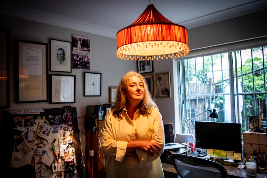 A woman stands arms crossed in a small study underneath an orange light shade, surrounded by bookshelves and a computer desk