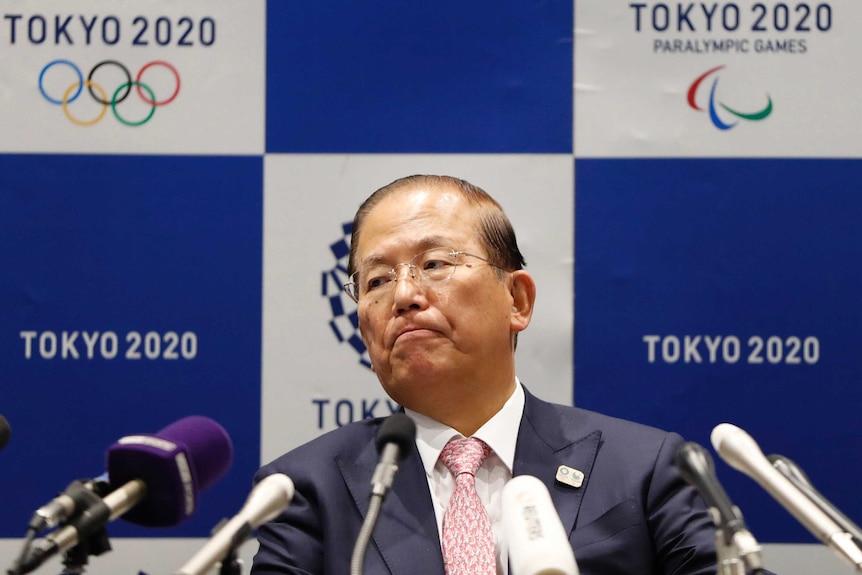 A serious looking Tokyo Olympics executive sits in front of microphones during a press conference.