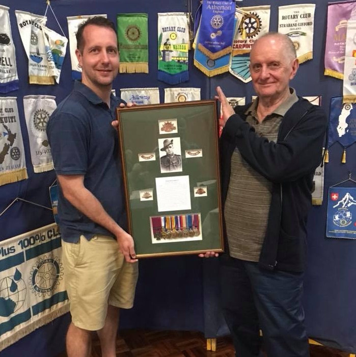 John and David Dalziel holding a commemorative display containing a photo, medals and text telling the story of Harry Dalziel.