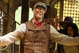 Dwayne Johnson smiles widely, his arms outstretched, standing inside an old-fashioned room with people celebrating behind him